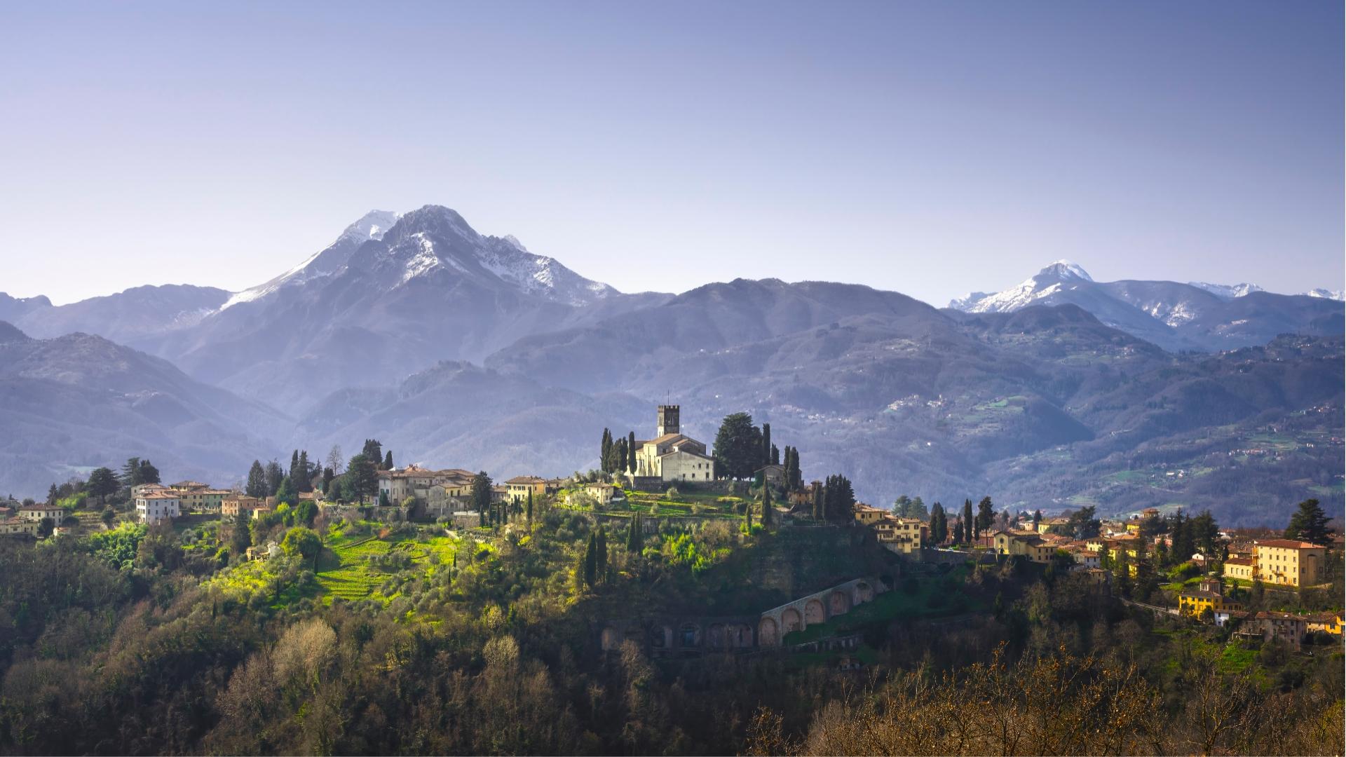 Paesaggio montano con villaggio e chiesa, circondato da colline verdi e montagne innevate.