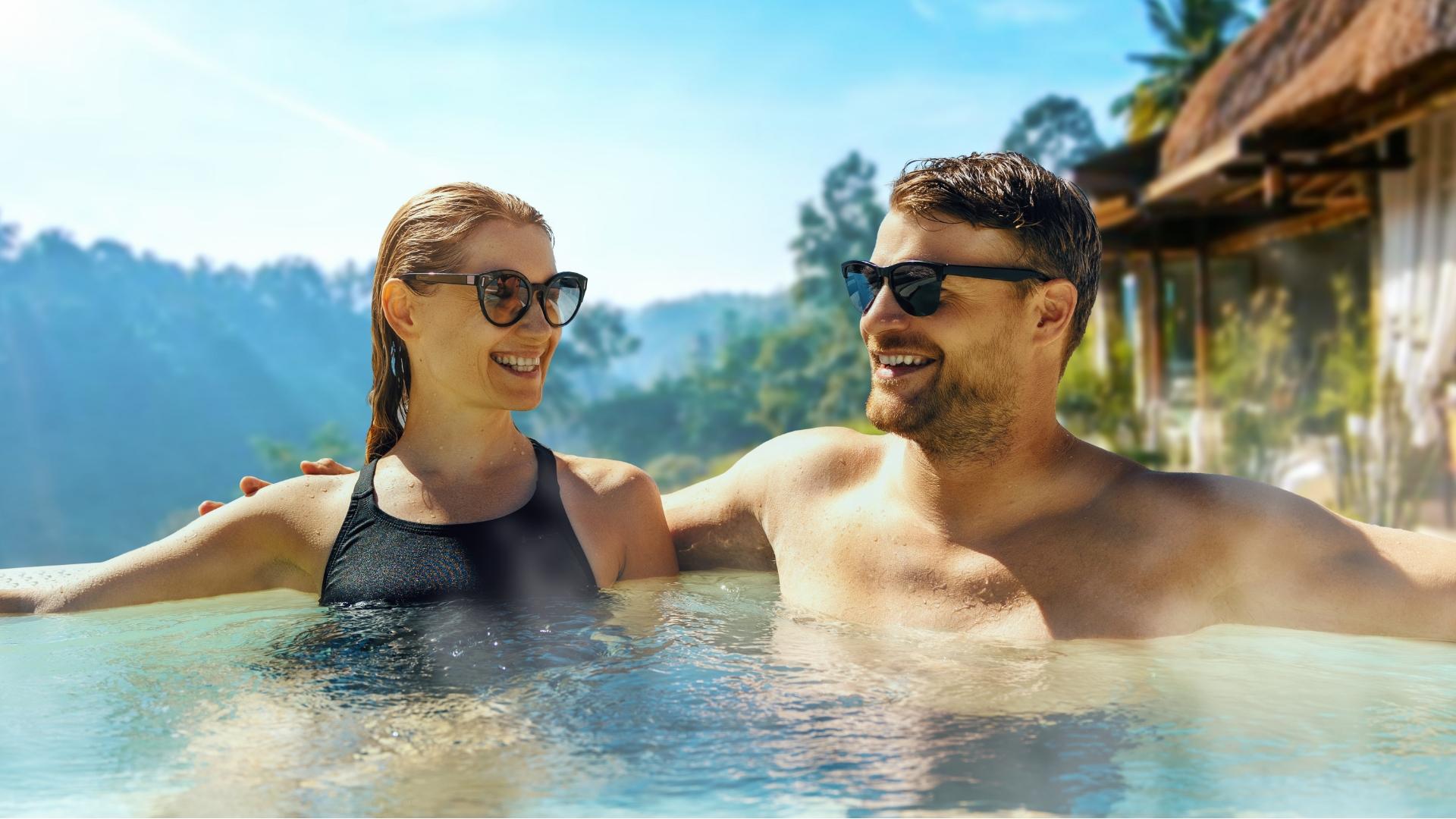 Coppia felice in piscina, indossano occhiali da sole, circondati da paesaggio tropicale.