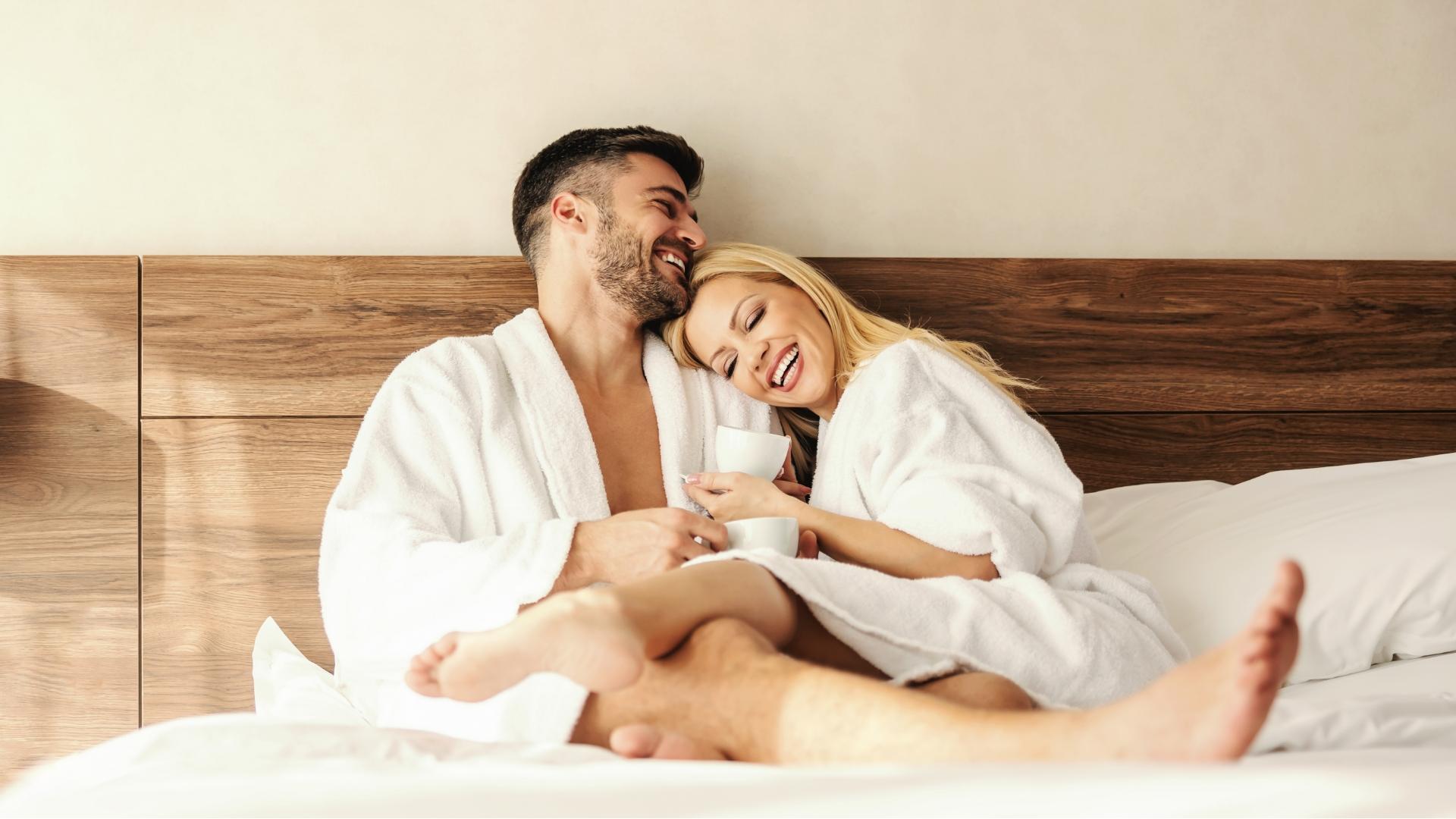 Coppia sorridente in accappatoio bianco, rilassata a letto con tazze da caffè.