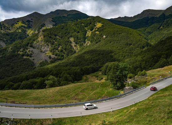 Strada tortuosa tra montagne verdi, due auto percorrono la curva sotto un cielo nuvoloso.