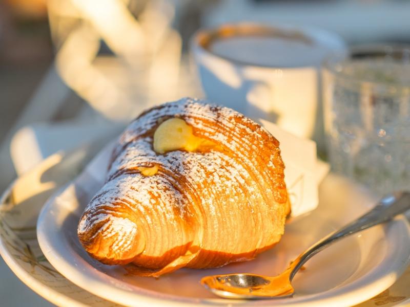 Cornetto con zucchero a velo su piatto, accanto a tazza di caffè.